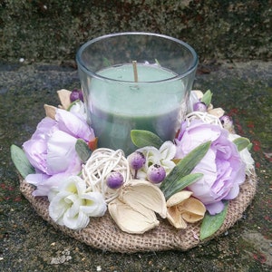 May include: A green candle in a glass jar sits on a burlap base decorated with purple and white flowers, dried flowers, and wooden balls.