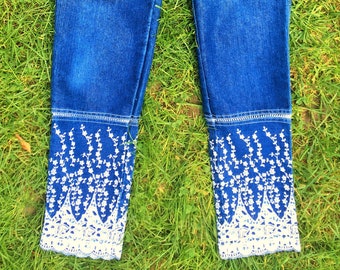 May include: Blue denim jeans with white embroidered floral trim at the bottom of the legs.