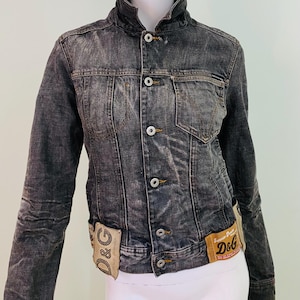 Puede incluir: Chaqueta vaquera negra descolorida con cuello alto y cierre de botones. Cuenta con dos bolsillos delanteros y un pequeño bolsillo en el pecho. Incluye etiquetas de la marca D&G en la parte inferior delantera. La chaqueta se muestra en un maniquí.