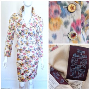 Puede incluir: Un traje de falda blanco con estampado floral y una chaqueta corta. La chaqueta tiene un cuello ancho y botones dorados. La falda es de largo hasta la rodilla y tiene una silueta ajustada. El traje está etiquetado con "John Galliano" y "Paris".