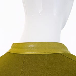 以下が含まれることがあります： コントラストの効いたシャルトルーズレザーの襟が付いたグリーンの衣服のクローズアップ。襟はオリーブグリーンの生地に縫い付けられています。白いマネキンに飾られた衣服。