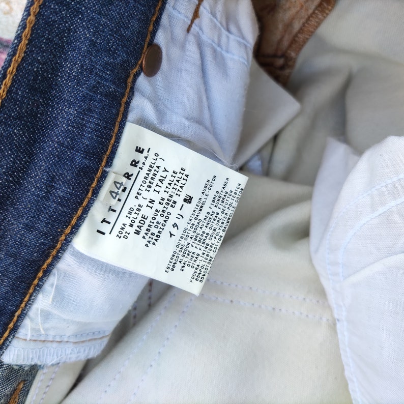 May include: A white tag with black text attached to a pair of blue jeans. The tag reads "ITT44RR S.P.A. ZONA IND. PETTORANELLO DI MOLISE (ISERNIA) MADE IN ITALY FABRIQUE EN ITALIE PAIS DE ORIGEN: ITALIE FABRICADO EM ITALIA イタリー製".