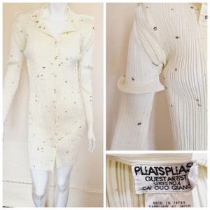 Puede incluir: Vestido blanco de punto de canalé con botones en la parte delantera y diseño plisado. El vestido tiene una etiqueta que dice "PLEATSPLIAS GUEST ARTIST SERIES NO.4 CAI GUO QIANG MADE IN JAPAN FABRIQUE AU JAPON".