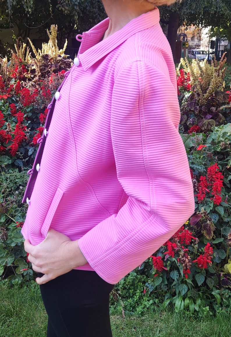 May include: A pink ribbed jacket with white buttons and a long sleeve. The jacket is made of a soft, stretchy material and has a relaxed fit.