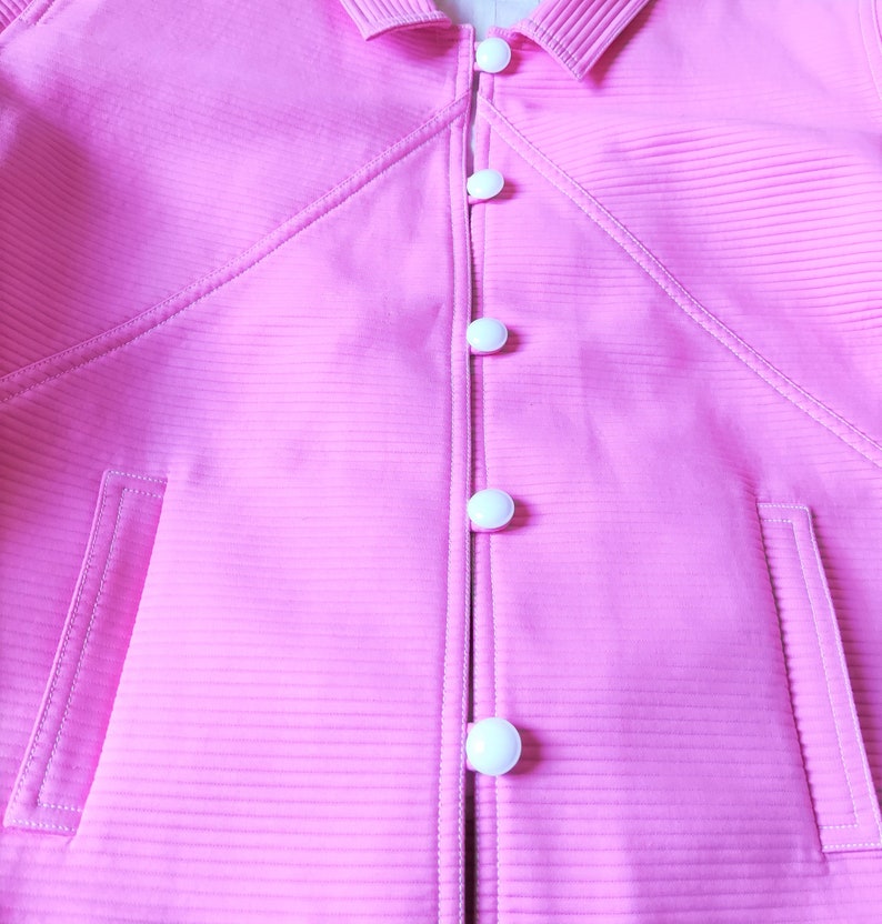 May include: A pink jacket with a ribbed texture and a row of white buttons down the front.