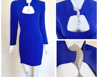 May include: A royal blue long-sleeved dress with a keyhole neckline and a jeweled embellishment at the collar. The dress has a fitted silhouette and a straight hemline.