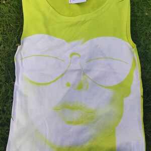 May include: A lime green sleeveless tank top with a white graphic print of a woman's face wearing sunglasses. The graphic is in a retro style with a bold outline. The text "courr&egrave;ges" is printed on the top in black.