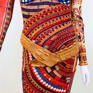 Op de afbeelding: Een jurk met lange mouwen met een levendig, veelkleurig patroon met rode, blauwe, oranje en gele tinten. Een lichtbruine, getextureerde riem accentueert de taille. De jurk heeft een uniek, artistiek ontwerp.