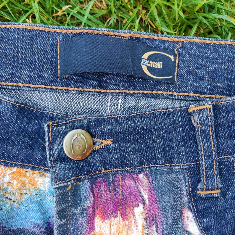 May include: Close-up of a pair of blue denim jeans with a colorful abstract print on the bottom. The jeans have a brown leather patch with the text "Just Cavalli" in white.