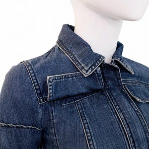 May include: A close-up of a denim jacket in a medium blue wash. The jacket features a classic collar, two chest pockets, and detailed stitching. The jacket is displayed on a white mannequin.