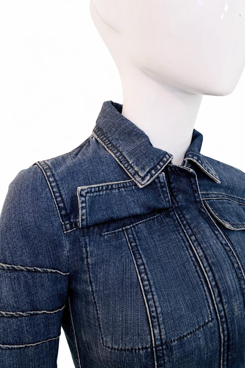 May include: A close-up of a denim jacket in a medium blue wash. The jacket features a classic collar, two chest pockets, and detailed stitching. The jacket is displayed on a white mannequin.