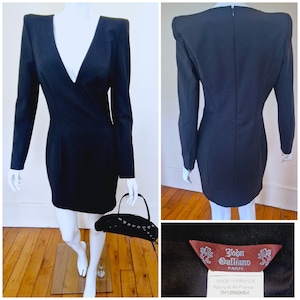 Puede incluir: Vestido negro mini con un escote en V profundo y mangas largas. El vestido tiene una silueta ajustada y presenta hombreras exageradas. La etiqueta dice "John Galliano Paris" y "Hecho en Francia, Fabrique en France, 7H12060054".