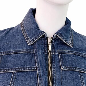 May include: A close-up of a blue denim jacket with a silver zipper. The jacket has a classic collar, two chest pockets, and visible stitching details. The jacket is displayed on a mannequin.