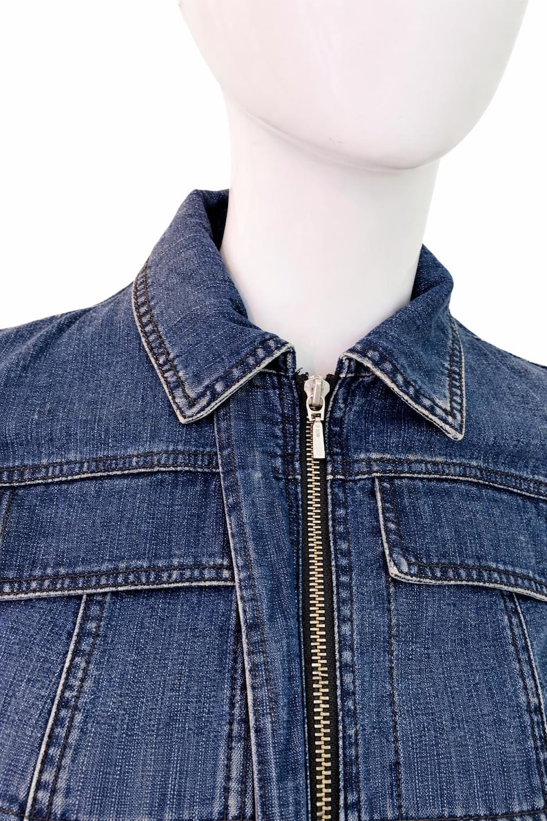 May include: A close-up of a blue denim jacket with a silver zipper. The jacket has a classic collar, two chest pockets, and visible stitching details. The jacket is displayed on a mannequin.