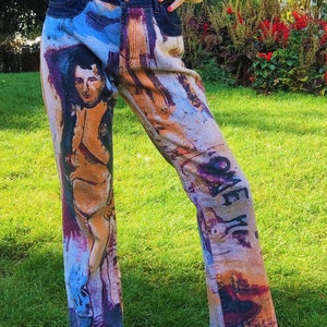 May include: Blue denim jeans with a colorful abstract print featuring a portrait of a person. The print includes a variety of colors, including blue, orange, yellow, and purple.