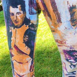 May include: A pair of pants with a colorful, abstract print featuring a portrait of a man in a military uniform. The print includes shades of blue, purple, orange, and white.