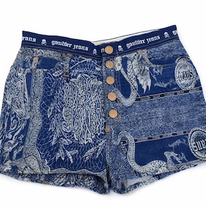 May include: Blue denim shorts with a unique pattern featuring ornate designs and animal figures. The shorts have a high waist, button closure, and the text "Gaultier Jeans" printed on the waistband.