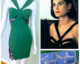 Puede incluir: Un minivestido verde con detalles de encaje negro y cortes. El vestido se exhibe en un maniquí. Se incluye una foto de una mujer con un vestido negro similar. Una etiqueta dice "Thierry Mugler PARIS".