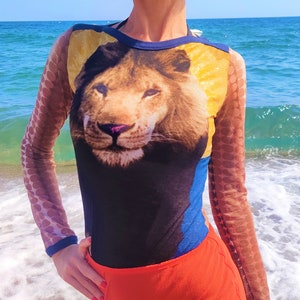 May include: A woman wearing a black, blue, and yellow long-sleeved shirt with a lion's face printed on the front. The shirt has a sheer, brown, polka-dot pattern on the sleeves. She is wearing a bright orange skirt.