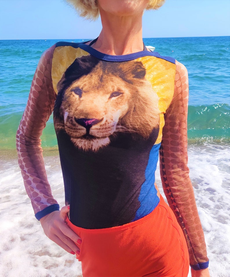 May include: A woman wearing a black, blue, and yellow long-sleeved shirt with a lion's face printed on the front. The shirt has a sheer, brown, polka-dot pattern on the sleeves. She is wearing a bright orange skirt.