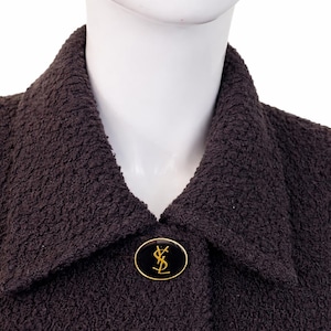 May include: A dark brown textured jacket with a pointed collar and a single circular button closure. The button is black with a gold-toned YSL logo in the center, surrounded by a thin gold-toned rim, creating a distinct focal point on the garment.