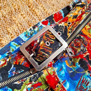 May include: Close-up of a silver belt buckle with the text "J.G. Jeans" on it. The buckle is attached to a black fabric belt with a colorful, abstract print.