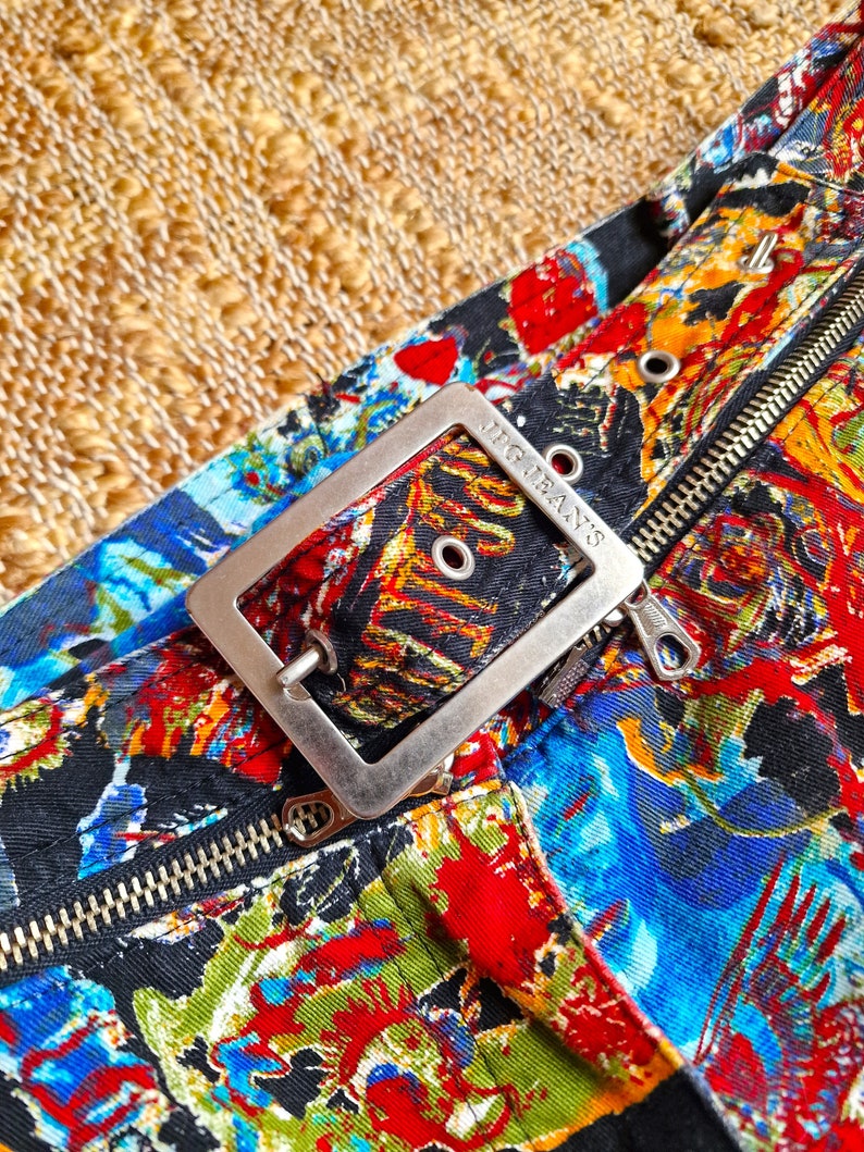 May include: Close-up of a silver belt buckle with the text "J.G. Jeans" on it. The buckle is attached to a black fabric belt with a colorful, abstract print.