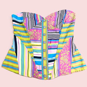 Può includere: Un corpetto patchwork colorato con scollatura a cuore. Il corpetto presenta un mix di motivi a righe e floreali nei toni del rosa, giallo, blu e nero. Ha un'abbottonatura frontale con bottoni argentati.