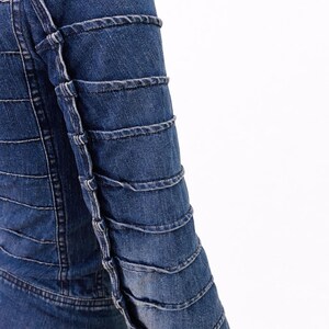 May include: A close-up of a denim jacket in a medium blue wash. The jacket features horizontal stitching across the body and decorative stitching on the sleeves. The sleeves have a layered, textured design.