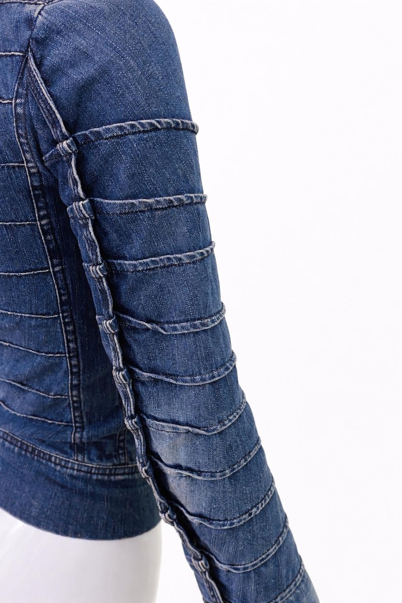 May include: A close-up of a denim jacket in a medium blue wash. The jacket features horizontal stitching across the body and decorative stitching on the sleeves. The sleeves have a layered, textured design.