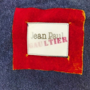 Könnte beinhalten: Ein rotes Quadrat mit gelbem Rand umschließt ein weißes Etikett mit dem Text "Jean Paul GAULTIER" in schwarzer und rosa Schrift.