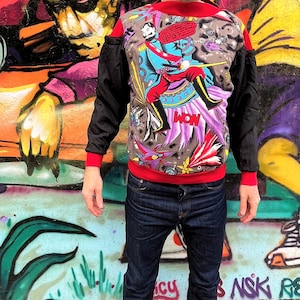 Könnte beinhalten: Ein schwarzes und rotes Sweatshirt mit einem bunten Grafikdruck eines Mannes, der ein lilafarbenes Wesen reitet. Die Grafik enthält das Wort "WOW" und eine Sprechblase mit Text. Das Sweatshirt hat ausgeschnittene Schultern und wird über einer blauen Jeans getragen.