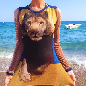 May include: A sheer, long-sleeved dress with a lion print. The dress is yellow, blue, and brown with a mesh fabric. The lion is facing forward with its mouth closed.