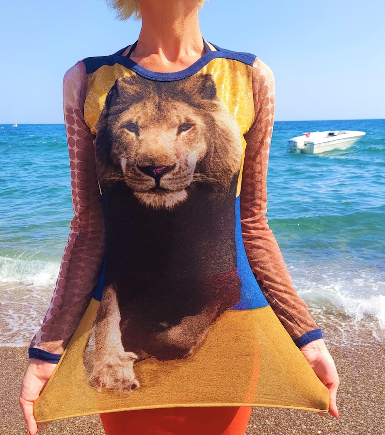 May include: A sheer, long-sleeved dress with a lion print. The dress is yellow, blue, and brown with a mesh fabric. The lion is facing forward with its mouth closed.