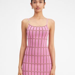 May include: A sleeveless mini dress in a vibrant pink hue featuring a structured grid pattern in a contrasting brown tone. The garment has thin spaghetti straps and a straight neckline, with a small gold-toned metal hardware detail on the right strap.