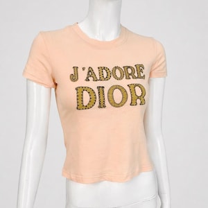 Può includere: Una t-shirt color pesca a maniche corte con la scritta "J'ADORE DIOR" in un carattere dorato e strutturato. La maglietta è esposta su un manichino. Ha un girocollo e un taglio corto.