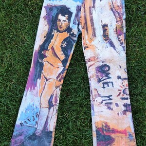 May include: A pair of blue denim jeans with a colorful, abstract print featuring a portrait of a man in a military uniform. The print also includes the words "Come Me" and "Daydream".