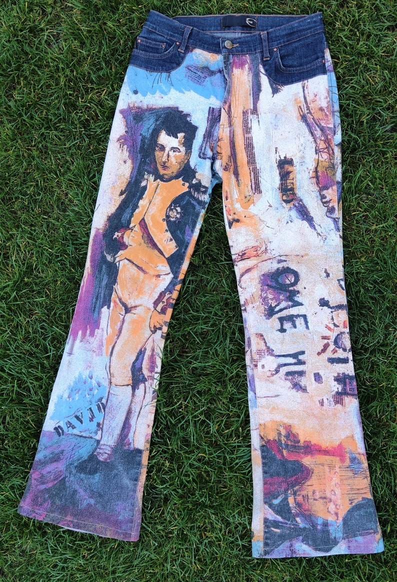 May include: A pair of blue denim jeans with a colorful, abstract print featuring a portrait of a man in a military uniform. The print also includes the words "Come Me" and "Daydream".