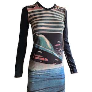 Puede incluir: Un vestido de manga larga con un diseño único. La parte superior presenta rayas horizontales en tonos grises y rosas, mientras que la parte inferior muestra un gráfico azul y negro. Las mangas son de color negro liso con costuras en contraste.