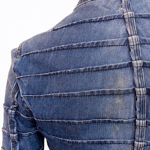 May include: Close-up of a blue denim jacket with horizontal and vertical stitched detailing. The jacket has a structured design with a collar and a tailored fit. The stitching is a lighter shade of blue, creating a contrast.