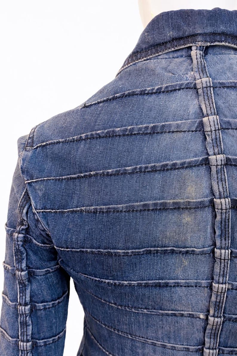 May include: Close-up of a blue denim jacket with horizontal and vertical stitched detailing. The jacket has a structured design with a collar and a tailored fit. The stitching is a lighter shade of blue, creating a contrast.