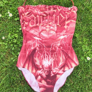 May include: Red one-piece swimsuit with a graphic print of a demon with wings and lightning bolts. The swimsuit has a halter neck strap.