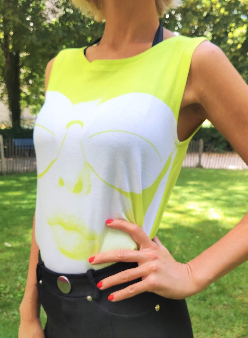 May include: A sleeveless lime green tank top with a white graphic of a woman's face wearing sunglasses. The graphic is a stencil design with a yellow outline.