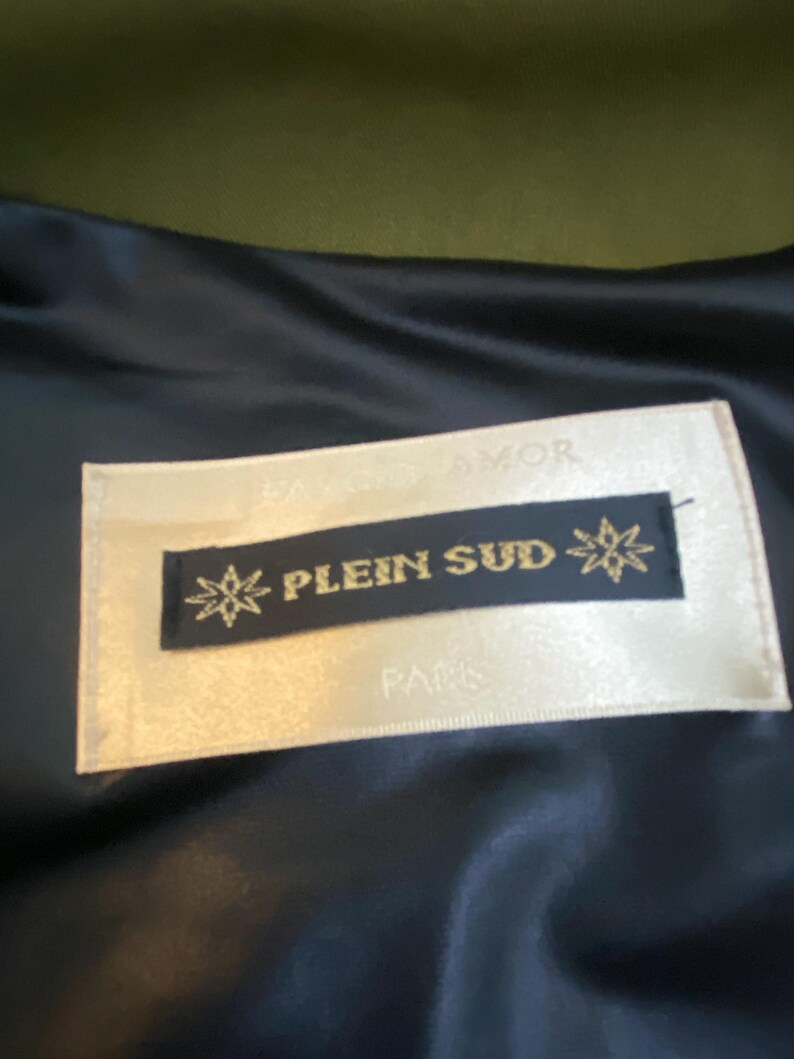 以下が含まれることがあります： 金色の文字で「PLEIN SUD」と書かれたラベルのクローズアップ。黒い背景にスターバーストのデザインがあります。ラベルはクリーム色の背景にあり、下に「PARIS」という文字があります。