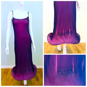 Puede incluir: Un vestido largo, sin mangas, de color púrpura con tirantes finos. El vestido tiene un dobladillo acampanado y una superposición transparente. El texto "00/81" y "RIGHT SIDE OVER-OUTSIDE" está impreso en el dobladillo. El vestido se exhibe en un maniquí.