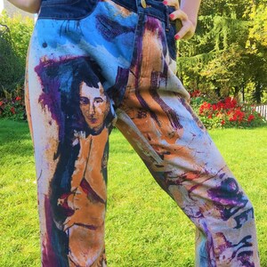 May include: A pair of blue denim jeans with a colorful abstract print featuring a portrait of a person. The print includes shades of blue, purple, orange, and brown.