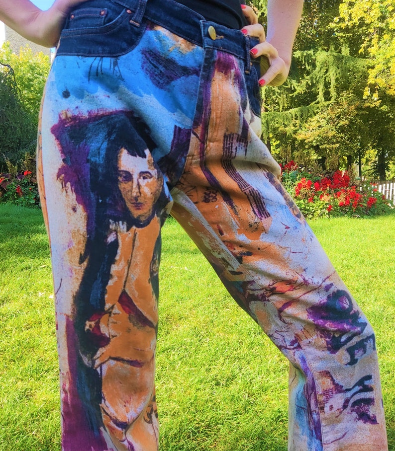 May include: A pair of blue denim jeans with a colorful abstract print featuring a portrait of a person. The print includes shades of blue, purple, orange, and brown.