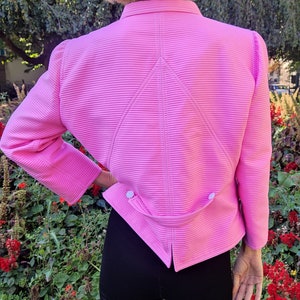 May include: A pink, long-sleeved, collared jacket with a ribbed texture and a pointed back design. The jacket has two white buttons on the back.