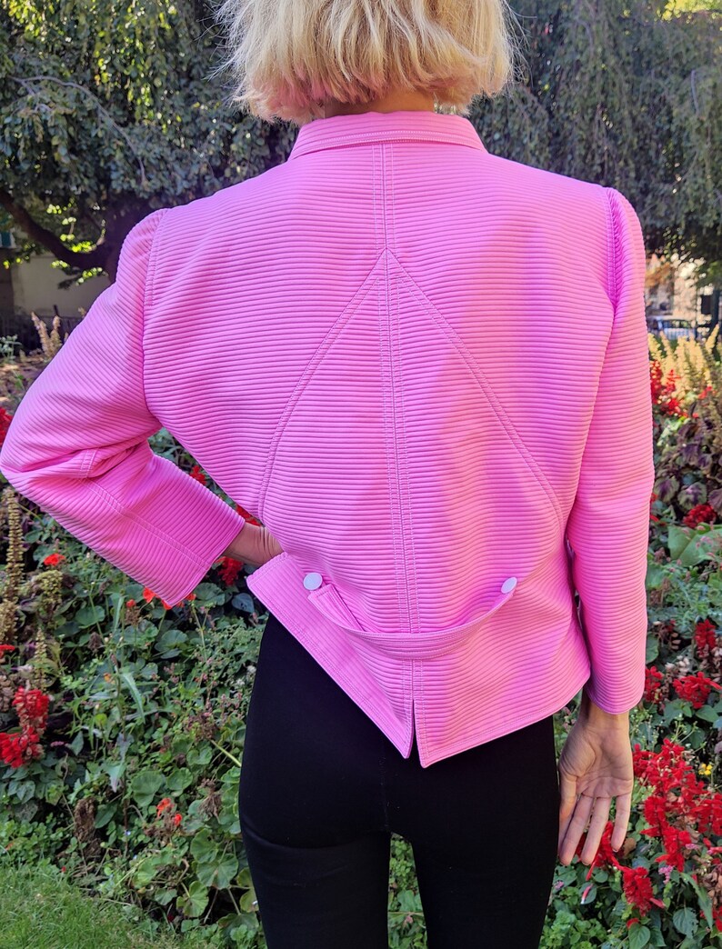 May include: A pink, long-sleeved, collared jacket with a ribbed texture and a pointed back design. The jacket has two white buttons on the back.