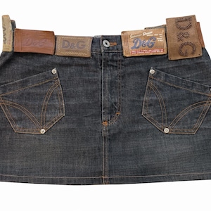 Puede incluir: Falda vaquera de lavado oscuro con trabillas y bolsillos de cuero marrón. La falda tiene dos bolsillos delanteros con costuras decorativas y remaches metálicos. Las trabillas llevan el logo "D&G".